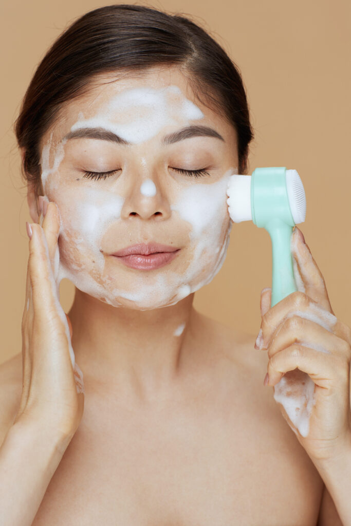 relaxed young asian female with massager washing face on beige background.