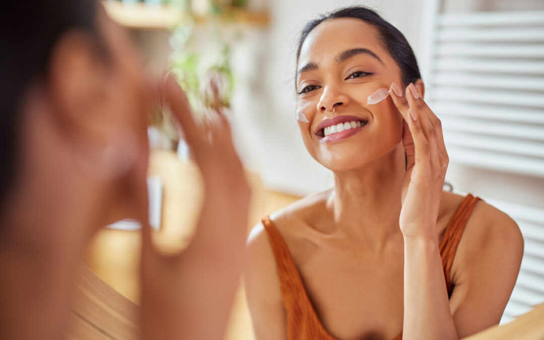 Portrait of smiling latin woman applying cream on her face while looking in mirror in her bathroom. Young multiethnic woman applying moisturizer on her cheek while looking in the mirror. Happy mexican girl using lotion every morning.