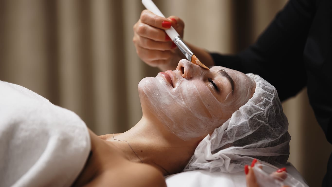 Close-up of a cosmetologist applying a cosmetic peeling mask to a client's face. The scene emphasizes the professional approach to skin care and the relaxing spa experience.